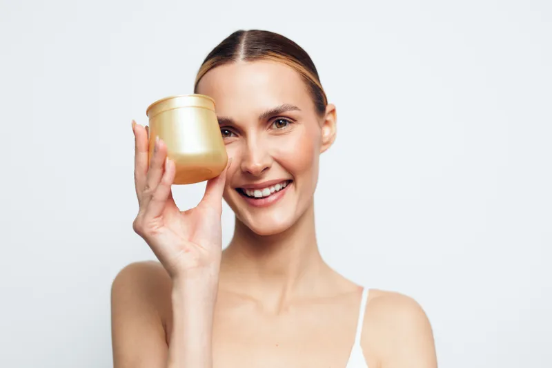 Smiling woman with clear skin holding cosmetic jar near face, natural makeup, beauty product concept, white background, healthy lifestyle, skincare routine, clean look