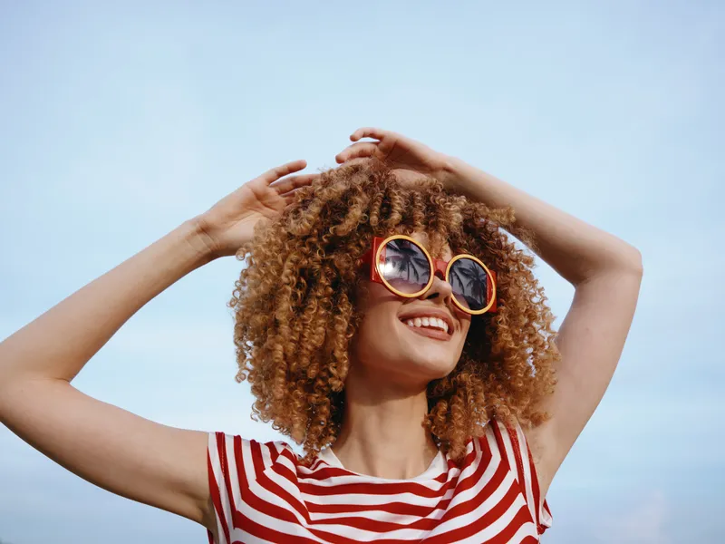 Portrait of a joyful woman with curly hair wearing round sunglasses and a red striped shirt, smiling broadly against a clear blue sky, sunny mood.