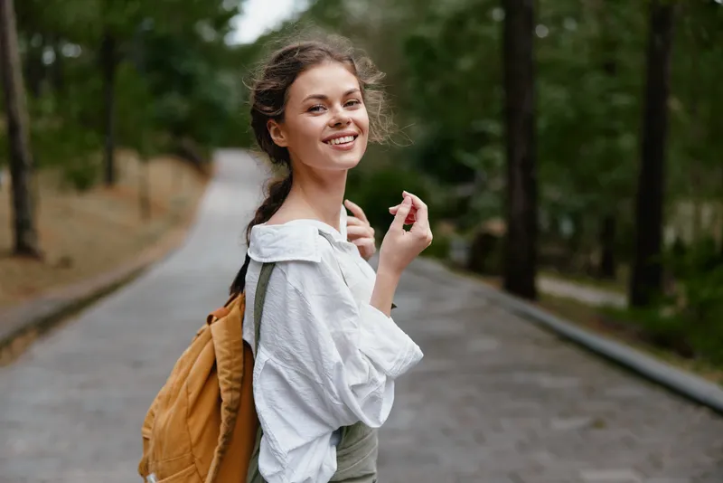 Young woman smiling in nature with a backpack, dressed in casual clothing, enjoying a carefree moment outdoors, perfect for travel and lifestyle themes