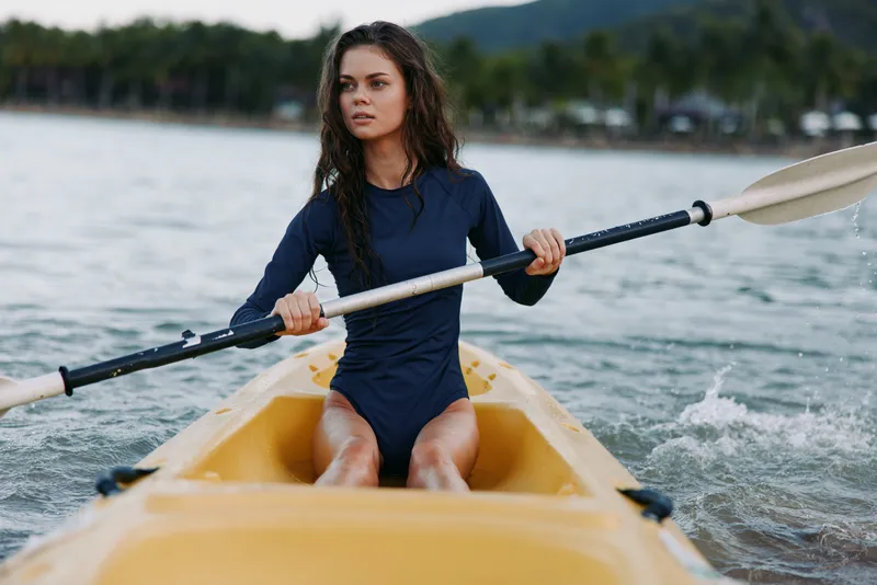 Dynamic outdoors scene of a focused female kayaker paddling through calm water in a yellow kayak, wearing a dark wetsuit, demonstrating balance, strength, and concentration.
