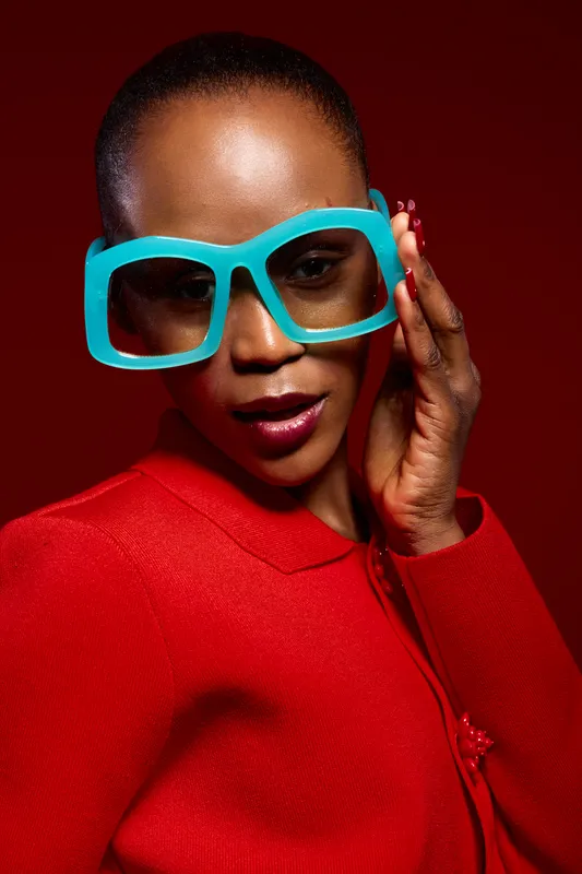 Close-up portrait of confident black woman with dark skin wearing large turquoise glasses and red sweater. Fashion concept with bold colors and stylish accessories on red background.