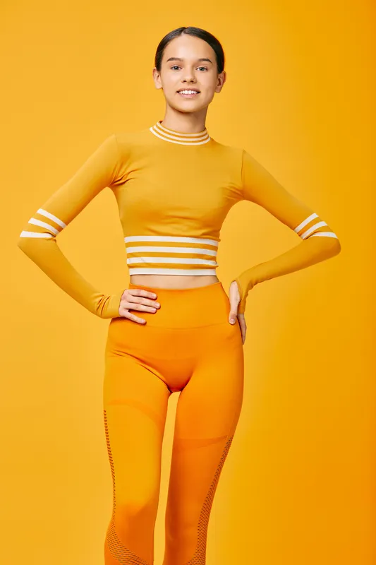 Fitness minded woman in bright orange athletic outfit posing with hands on hips in a studio, showcasing confidence, athleticwear style and energetic sport vibe against a bold orange background.
