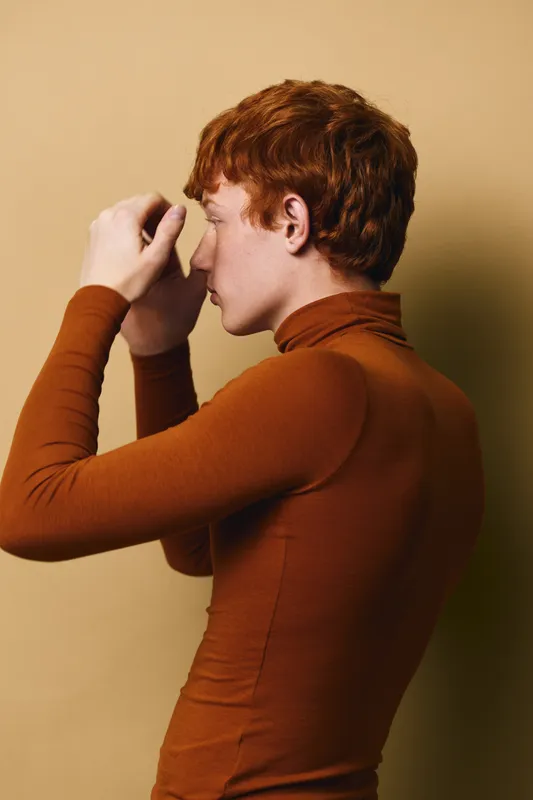 Redhead man with curly hair wearing brown turtleneck sweater standing sideways against beige background in thoughtful pose, portrait of young male model in studio lighting.