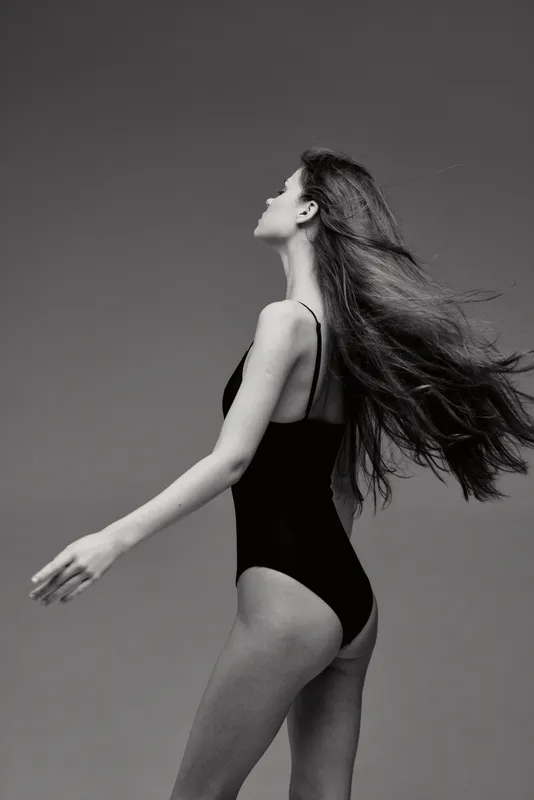 Black and white portrait of a woman with long hair wearing a black swimsuit against a plain background.