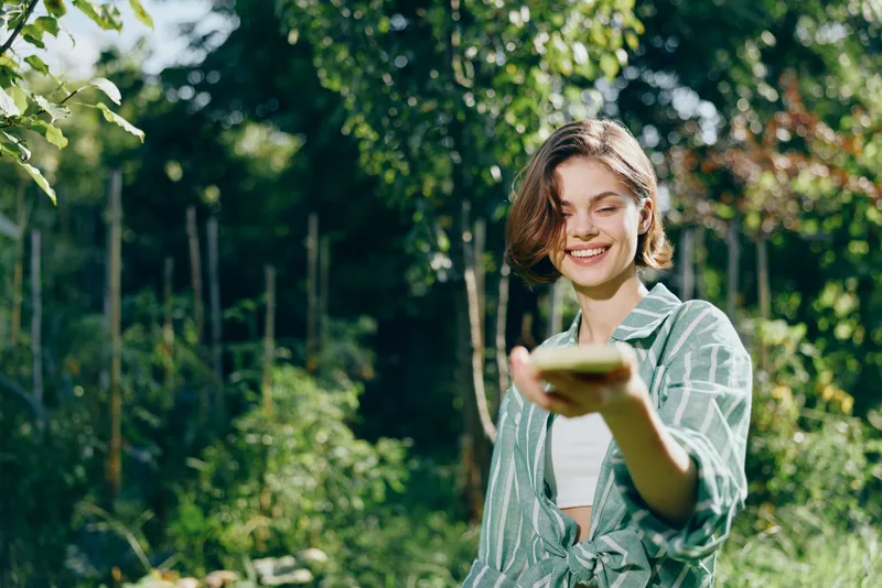Woman in casual green striped shirt stands outdoors in a sunny park, smiling as she lifts a phone toward the camera, inviting connection and friendly interaction.
