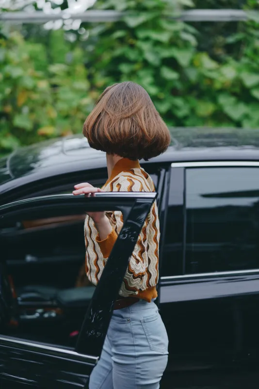 A woman with a short bob hairstyle steps toward a dark car, gripping the door as lush greenery surrounds the scene.