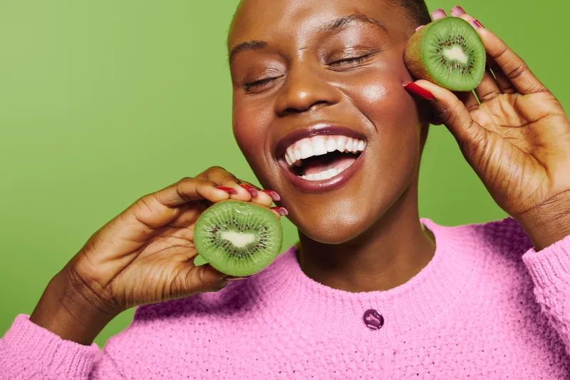 Happy African woman with dark skin holds fresh kiwi halves near face, smiling with closed eyes on green background. Concept of healthy lifestyle, fruit, wellness, and natural beauty.