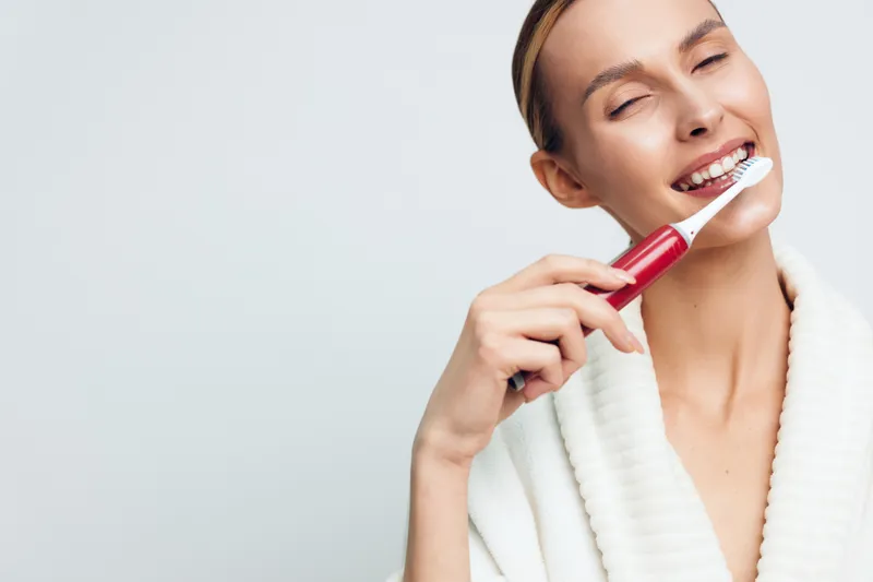 Young woman in white bathrobe brushing teeth with electric toothbrush on light background. Concept of dental care, hygiene, and morning routine with natural lighting and happy expression.