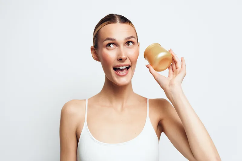 A happy young woman in a white tank top holds a jar of cream near her face while looking up with excitement. The bright studio background highlights skin care and beauty concepts.