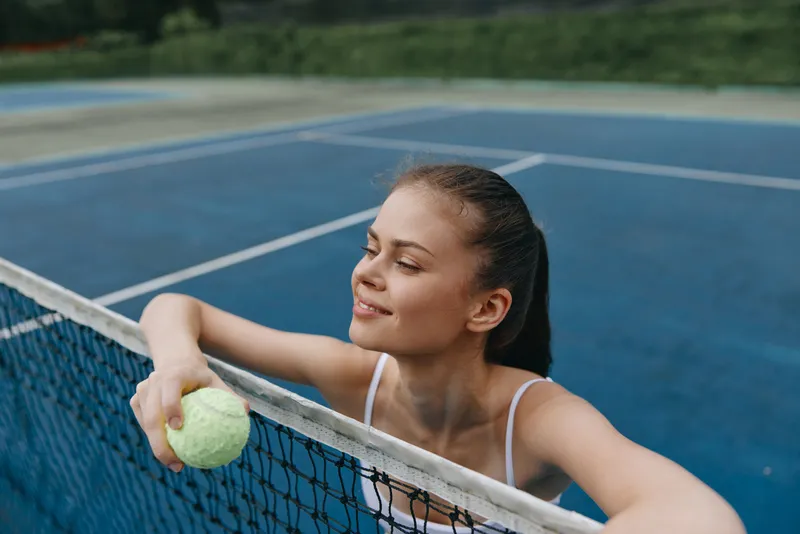 Young female athlete smiling with a tennis ball on a blue tennis court, showcasing determination and joy in sports, perfect for healthy lifestyle concepts