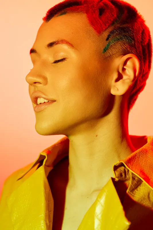 Woman with closed eyes and creative colorful hairstyle in vibrant lighting, wearing a yellow jacket, expressing calm and artistic mood.