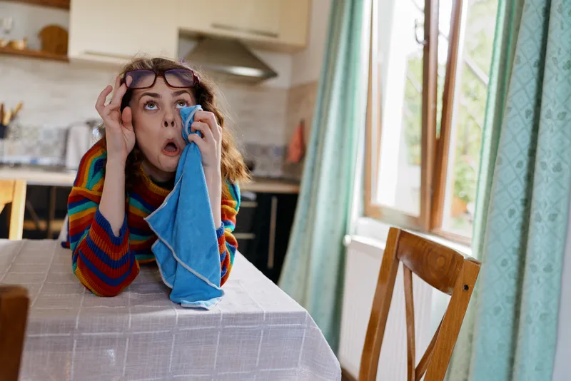 Woman in a striped sweater sits at a kitchen table, wiping her eye with a blue cloth, surprised expression. A candid home lifestyle moment focused on daily routine and self care.
