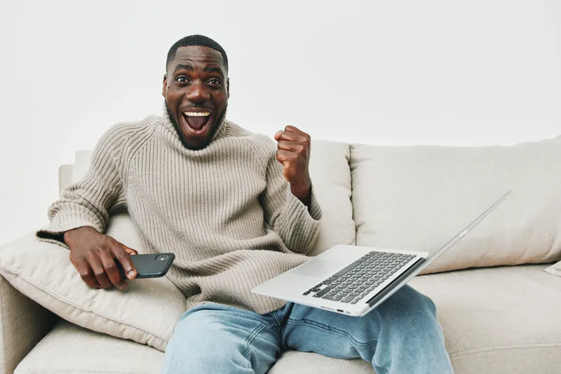 Cheerful young man celebrating success while working on a laptop at home, wearing a cozy sweater in a minimalist living room setting