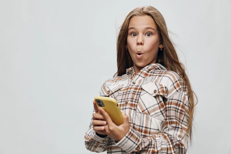Girl with long hair in plaid shirt looking surprised while holding yellow smartphone with both hands on gray background for communication and technology concept.