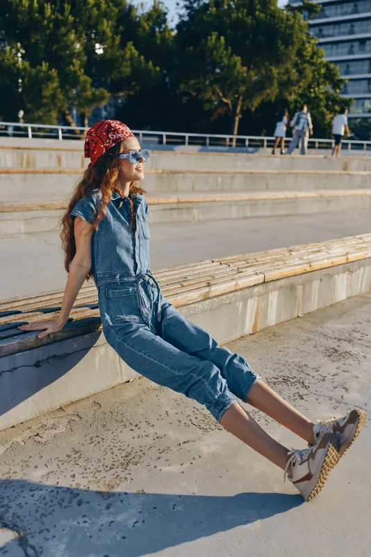 Lifestyle woman in modern boho-western denim street style sitting outdoors wearing bandana and sunglasses with film color tones in urban summer setting.