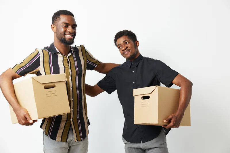 Two smiling African American men lifting cardboard boxes in a light and cheerful atmosphere, showcasing teamwork and friendship in a bright setting.