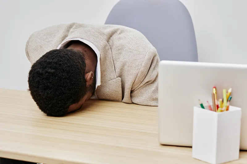 Frustrated young man with black hair leaning forward on a desk with a laptop and stationery, head resting on surface, expressing stress or exhaustion, in an office setting with neutral background. Peo