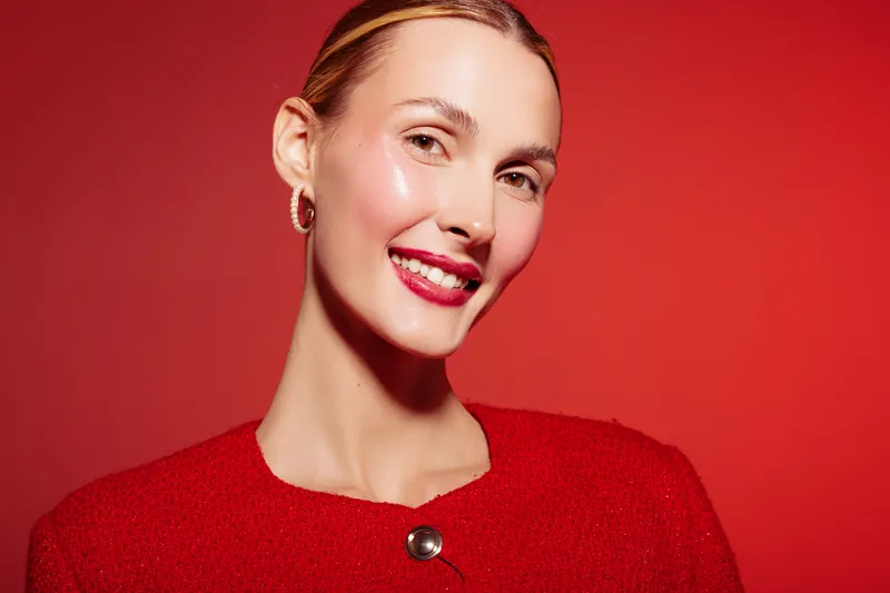 Smiling woman with shiny skin and red lipstick wears red outfit and gold earrings. Portrait on red background with studio lighting. Concept of beauty, fashion, and confident expression.