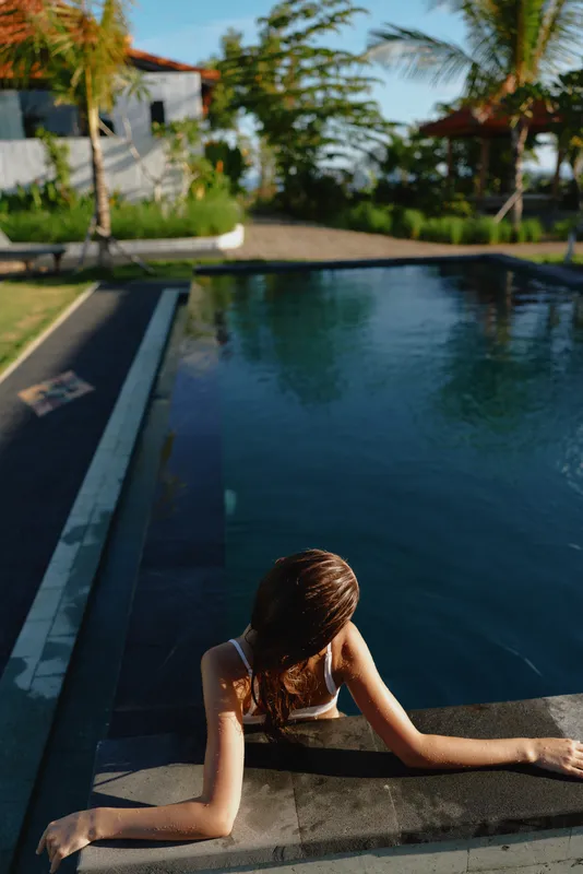 Young woman with wet hair leans on the pool edge as sunlight glints on the blue water, portraying a calm, intimate lifestyle moment by the water.