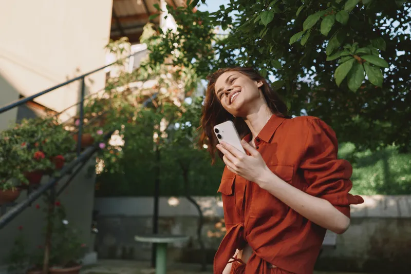 Cheerful woman outdoors wearing a red dress, smiling at a smartphone, sunlit garden backdrop, relaxed lifestyle moment, natural colors, soft breeze, candid and vibrant