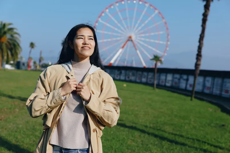 Woman in a casual jacket standing in an outdoor amusement park beside a ferris wheel on a sunny day, enjoying leisure, travel vibe, greenery, and a bright, relaxed smile.