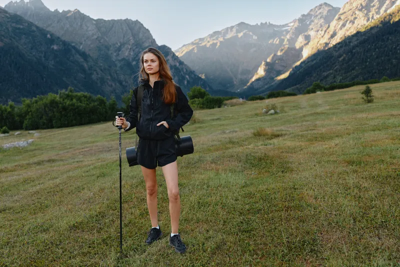 A cheerful hiker stands in a mountain meadow with trekking pole and camera gear, surrounded by towering peaks and clear sky, conveying adventure and outdoor exploration.