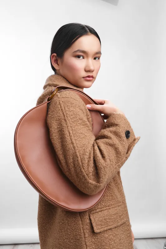 Asian woman stands in a modern studio, smiling softly while adjusting a stylish brown bag over her shoulder, exuding confidence and contemporary fashion appeal.