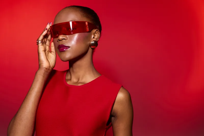 Elegant black woman with dark skin wearing futuristic red sunglasses and a red sleeveless dress poses against a red background. Concept for fashion, style, and modern beauty.