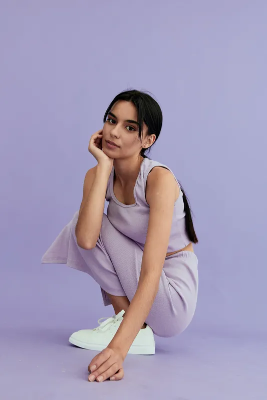 Young woman squatting indoors wearing casual lavender outfit and white sneakers against pale purple background in natural soft light.