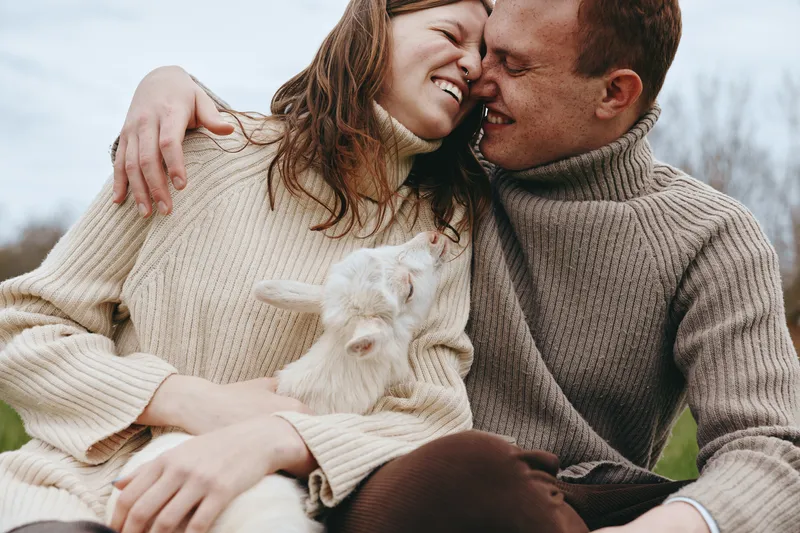Cheerful couple outdoors cuddling a goat kid, warm sweaters, relaxed pose, soft breeze, natural colors, rustic setting, sunny mood, candid affection, intimate moment and joyful laughter echoes in the