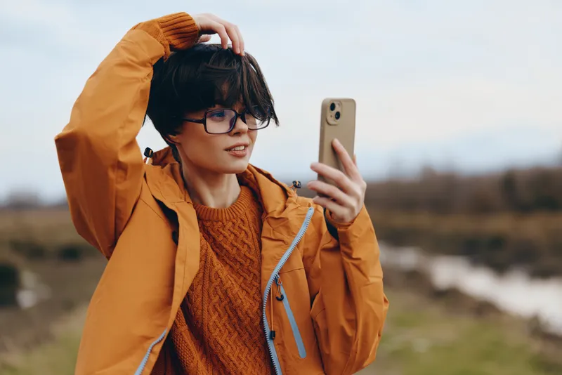 Woman lifestyle selfie smartphone outdoor jacket sweater glasses portrait, in orange coat she holds a phone for selfie, adjusts hair, soft daylight over coastal nature landscape, candid travel moment