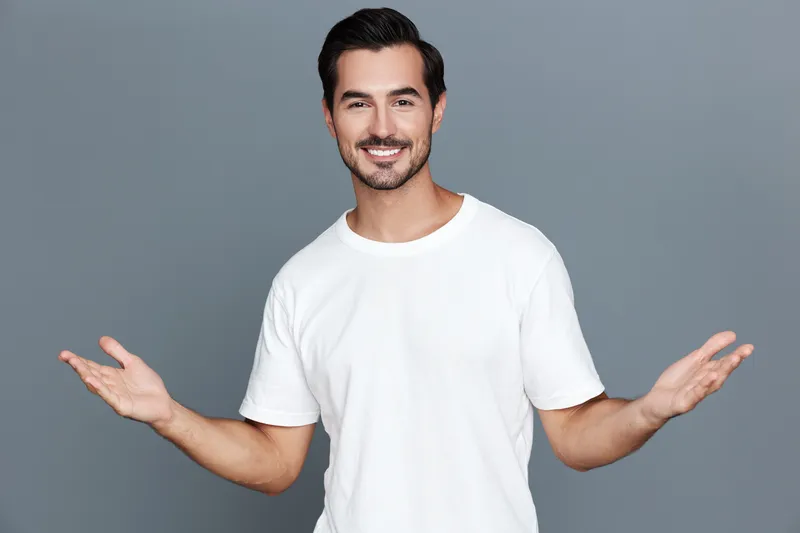 Charming smiling man wearing a simple white t-shirt stands with open arms, looking at the camera against a soft gray background. Positive vibes concept.
