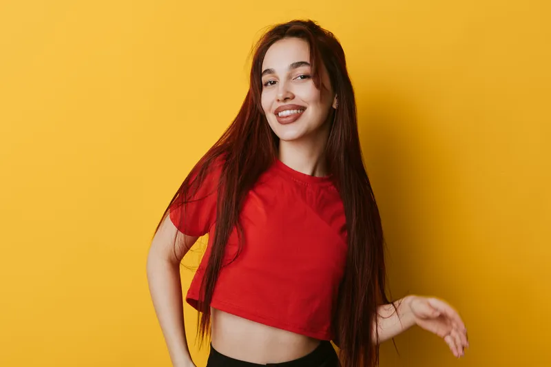 Young woman with long hair smiling and posing in red crop top against yellow background. Casual style, confident expression and relaxed posture for lifestyle and fashion portrait.
