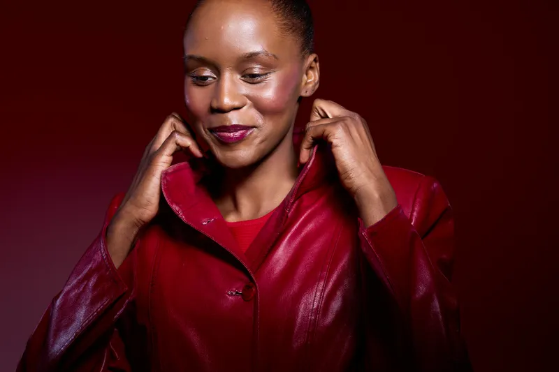 Confident African woman with dark skin wearing a red leather jacket adjusts the collar against a red background. Studio portrait with warm lighting and fashionable casual style.