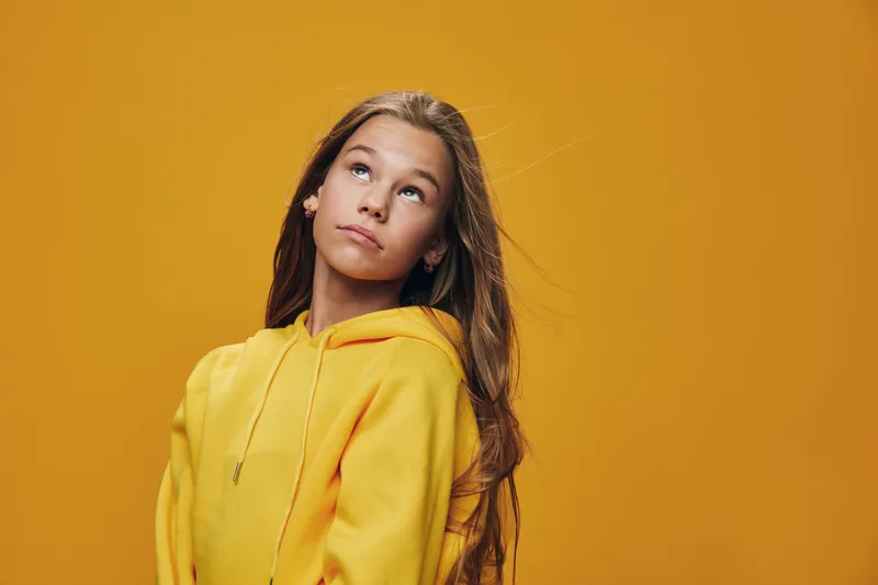 Girl in yellow hoodie looking up with thoughtful expression on orange background. Portrait of young female with long hair wearing casual clothing for fashion and lifestyle.