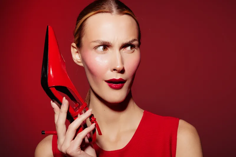 Woman in red dress holds red high heel shoe near face with curious expression. Bright red background and matching lipstick create bold fashion concept and style statement.