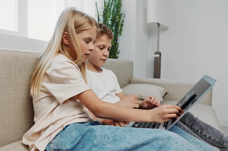 Two children sitting on a sofa using a laptop together in a bright room. Focused kids engaged in learning, technology, and digital education at home environment.