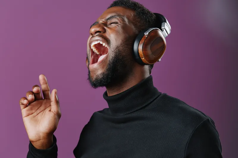 Energetic young man singing passionately with headphones on, wearing a black turtleneck, against a vibrant purple background, conveying joy and music engagement