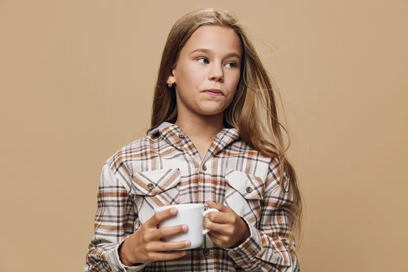 Girl wearing plaid shirt holds white mug with both hands looking to the side against beige background. Portrait of thoughtful young female with long hair in casual clothing indoors.
