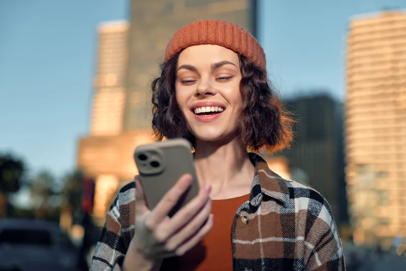 Woman with phone and bright smile in city at golden hour, wearing beanie and plaid jacket; candid lifestyle portrait with authenticity, golden hour glow, mindful living and emotional storytelling.