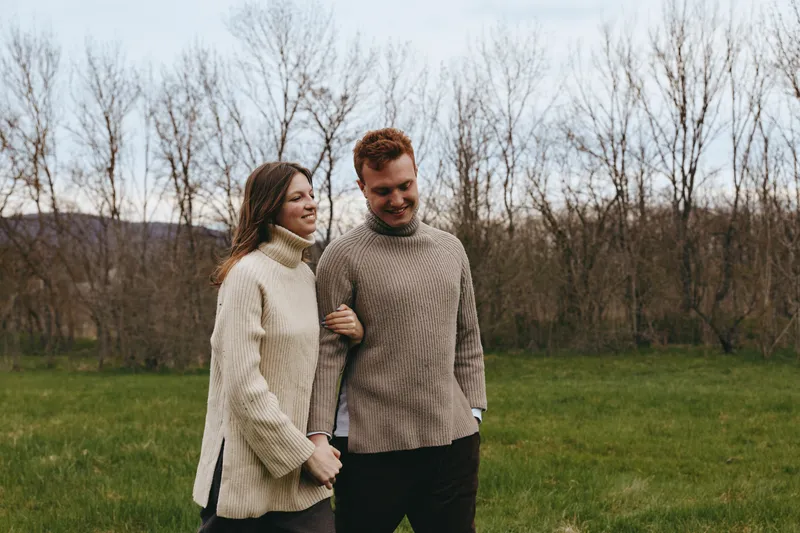 Couple walking hand in hand in an open field wearing chunky sweaters, conveying warmth and togetherness in a rustic landscape. Casual lifestyle scene highlighting companionship and natural light.
