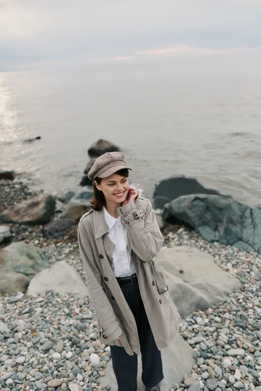 Young woman in beige coat and hat stands on rocky beach near calm sea, smiling and looking away. Concept for autumn fashion, casual style, and outdoor lifestyle by the water.