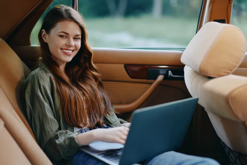 A young woman with long hair working on a laptop in a vintage car, smiling contently, reflecting a relaxed, inspirational mood amid nature.