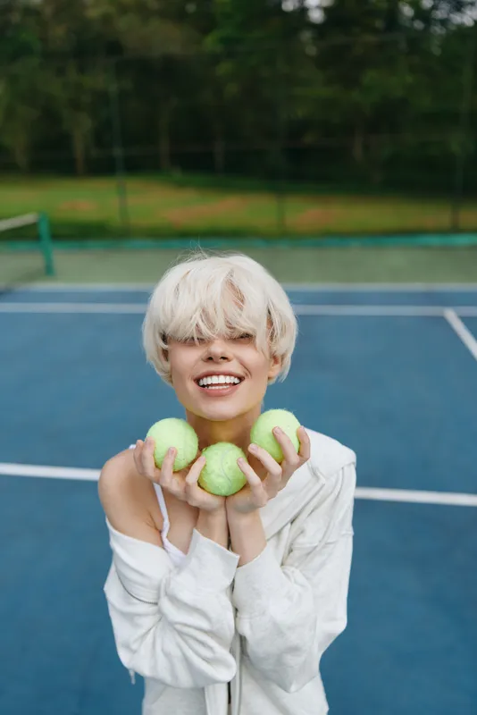 Young woman with short blonde hair smiles joyfully while holding green tennis balls on a blue court, showcasing a vibrant and energetic mood