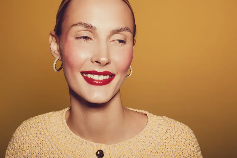 Smiling woman with red lipstick and hoop earrings wears beige knitted sweater on warm brown background. Close-up portrait with natural makeup and joyful expression. Beauty and fashion concept.