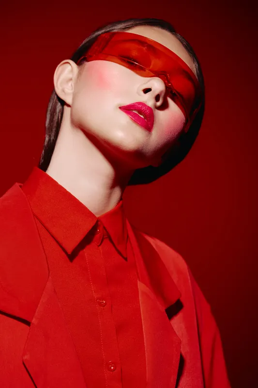 Fashion model wearing vibrant red clothing and futuristic orange glasses poses confidently against a red backdrop. Creative makeup and strong colors emphasize beauty concept.