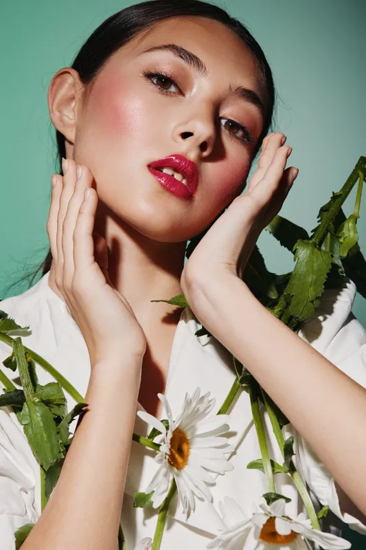 Fashion model with glossy red lips and flushed cheeks posing with white daisies. Portrait with soft lighting on a mint background. Concept for beauty, fashion, creative photography.