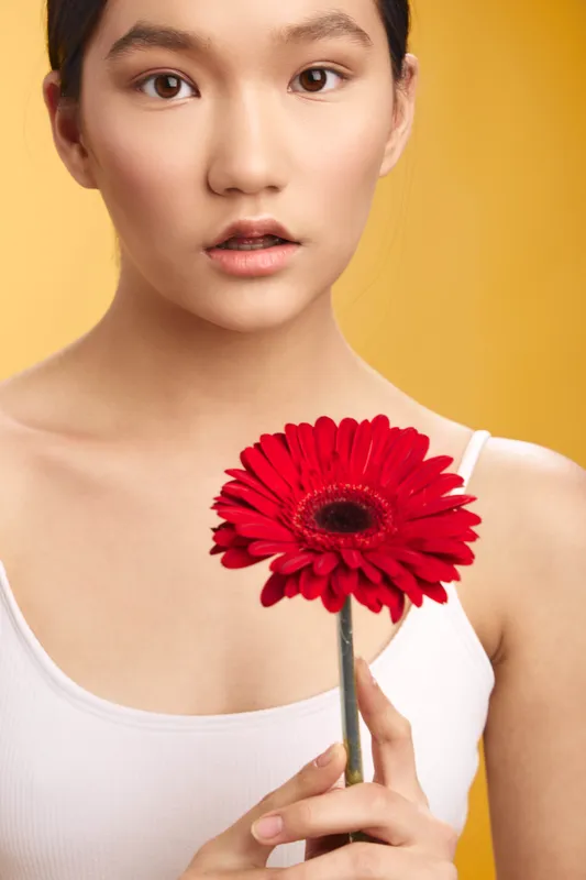 Asian woman holds a red flower against a warm yellow backdrop, a calm expression and soft lighting creating an elegant portrait that conveys beauty, focus, and gentle charm.