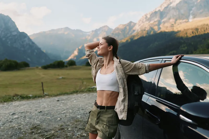 Young woman in casual clothes stands beside car in mountain landscape, enjoying fresh air and nature. Concept of lifestyle, travel, freedom, and outdoor adventure.