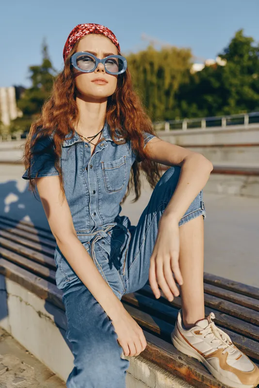 Lifestyle woman in modern boho-western denim street style wearing oversized blue glasses and red headband, sitting outdoors in film color warm sunlight.